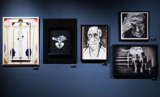 Una parete blu della mostra con cinque opere di autori diversi, ciascuna con la propria targhetta: a sinistra un’opera chiara in cornice bianca con una figura allungata al centro, poi una piccola opera su fondo nero, due ritratti in bianco e nero e una fotografia